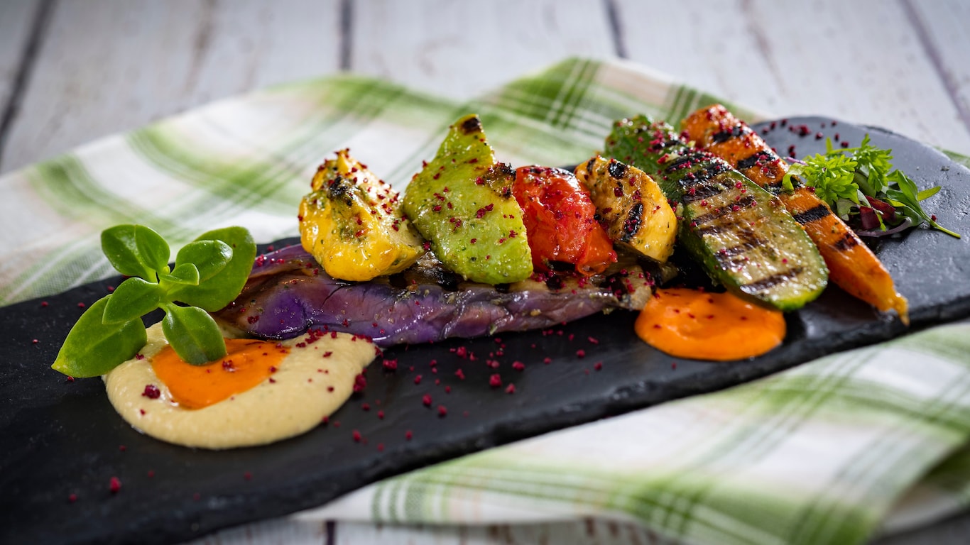 An assortment of grilled baby vegetables served with hummus cream and red pepper coulis, garnished with micro greens
