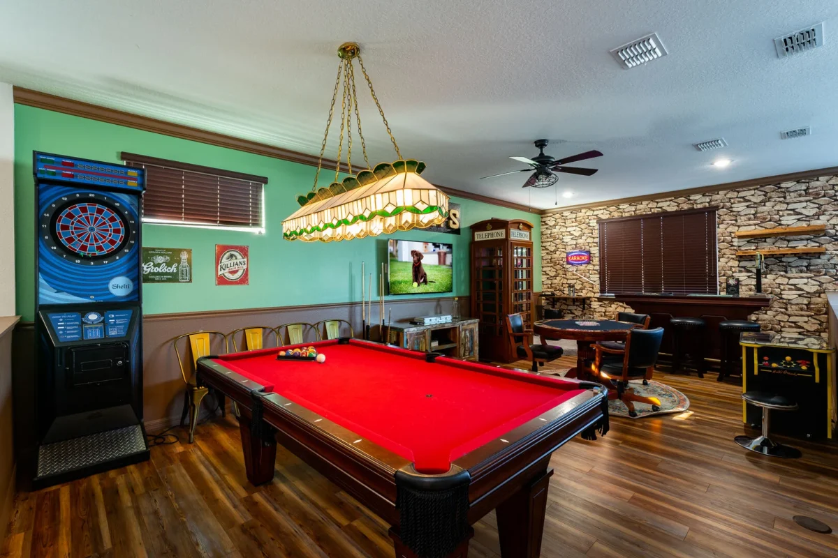 A spacious game room with red felt pool table, dartboard, bar seating, and entertainment area.