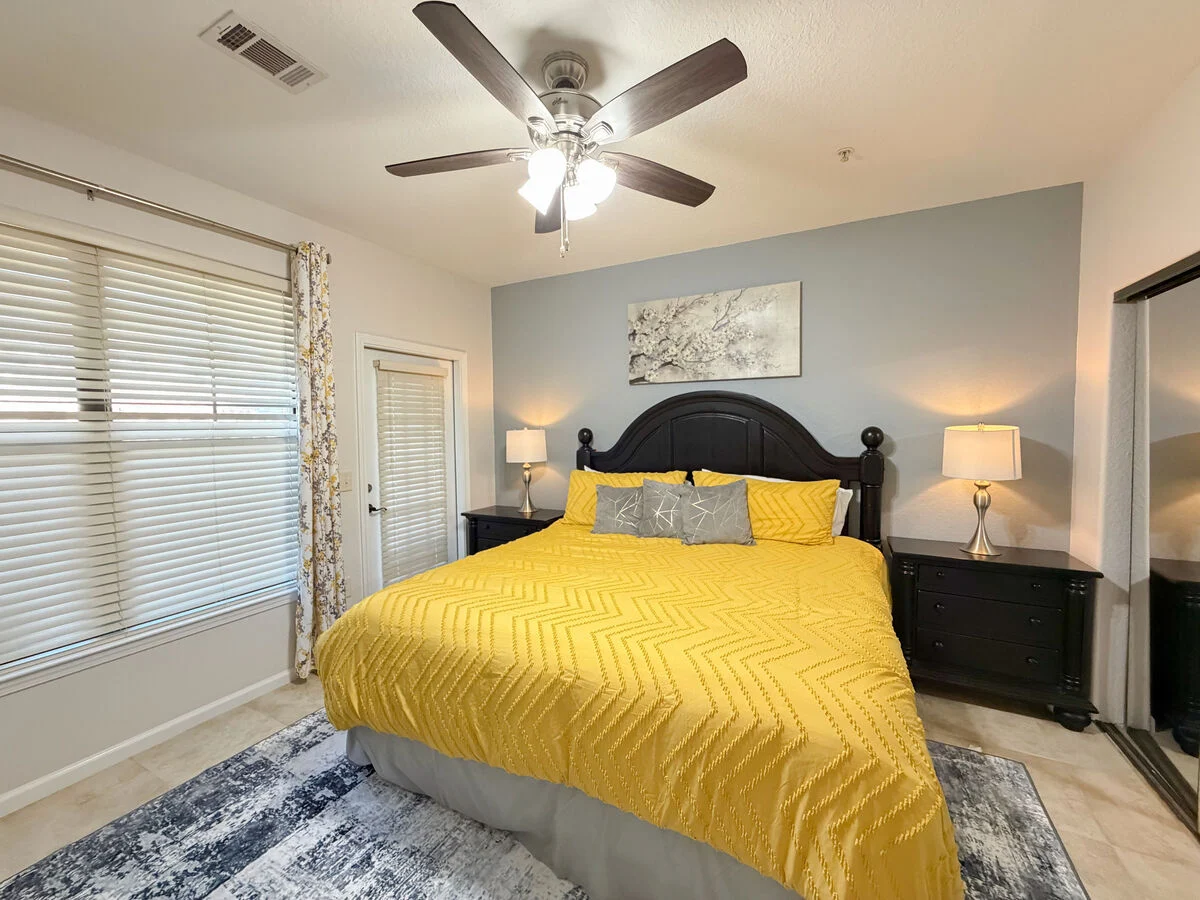 Bright bedroom with king bed, yellow bedding, ceiling fan, bedside lamps, and natural light from window. 