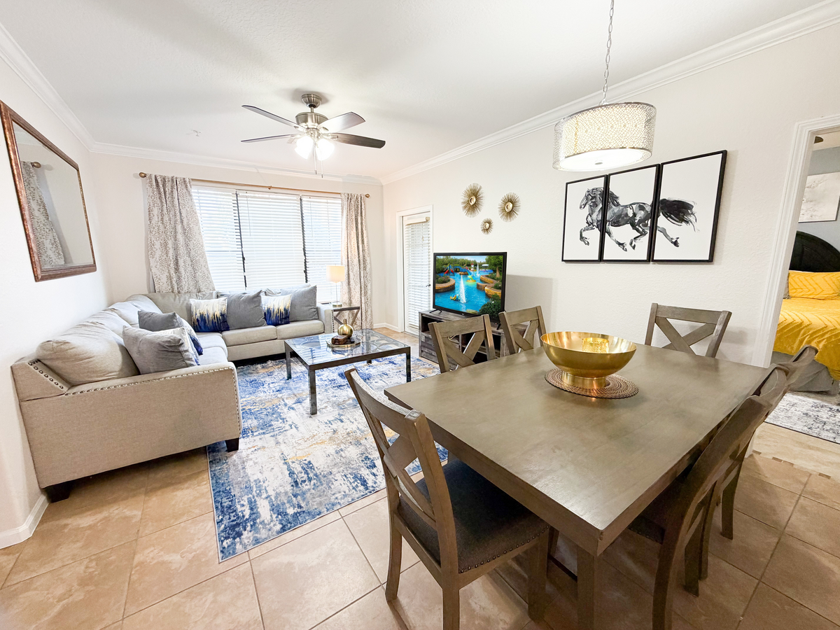 Open plan living and dining area with sectional sofa, dining table, TV, and natural light from large windows. 